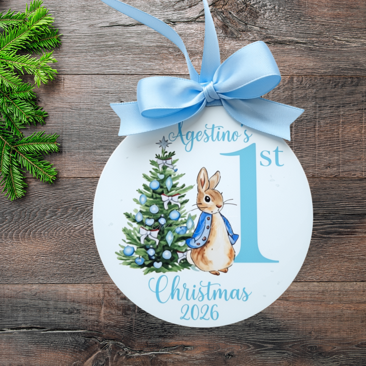 A round white Christmas ornament with a light blue ribbon bow, featuring a watercolor illustration of Peter Rabbit next to a decorated Christmas tree and the text 'Agostino's 1st Christmas 2026' on a rustic wooden background.