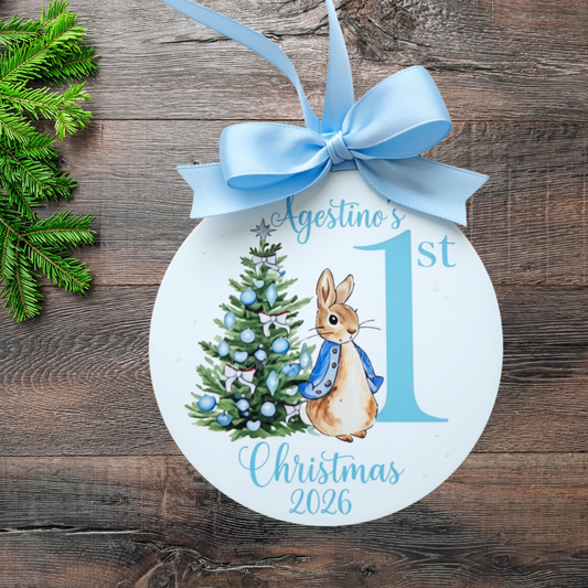 A round white Christmas ornament with a light blue ribbon bow, featuring a watercolor illustration of Peter Rabbit next to a decorated Christmas tree and the text 'Agostino's 1st Christmas 2026' on a rustic wooden background.
